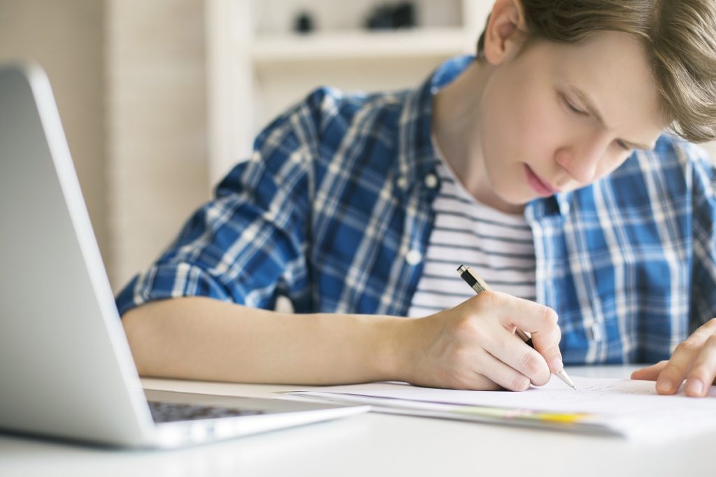 Boy writing on paper with his laptop open